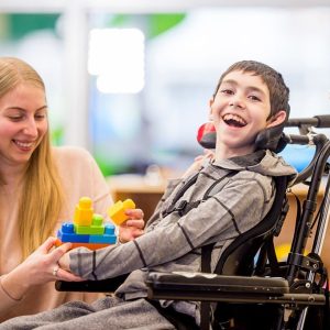 A caregiver is helping a young boy with a physical disability play with plastic block toys.